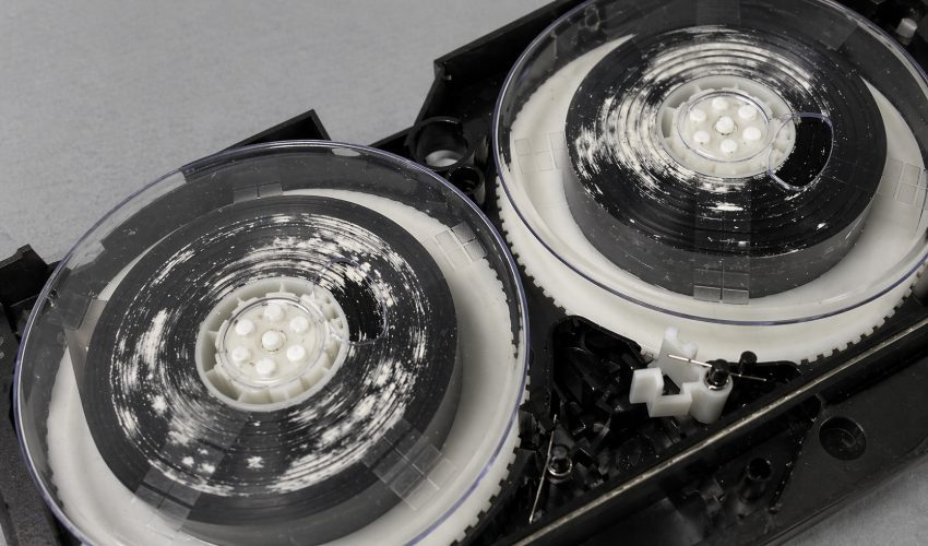 Close-up inside a VHS cassette showing white fuzzy mold growth around the reel hubs and on the magnetic tape ribbon, a classic sign of long-term storage in humid conditions