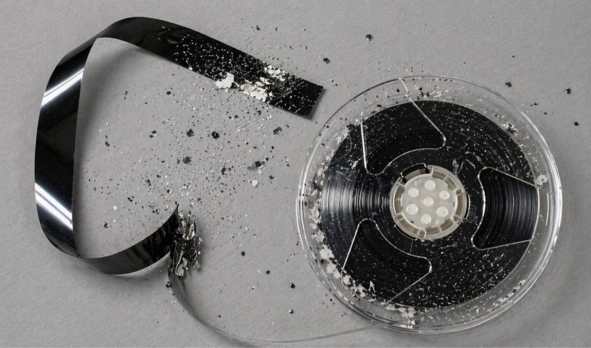 A VHS reel with magnetic tape ribbon and visible oxide particles shedding off the surface, showing the granular residue that builds up on playback heads when sticky shed syndrome sets in