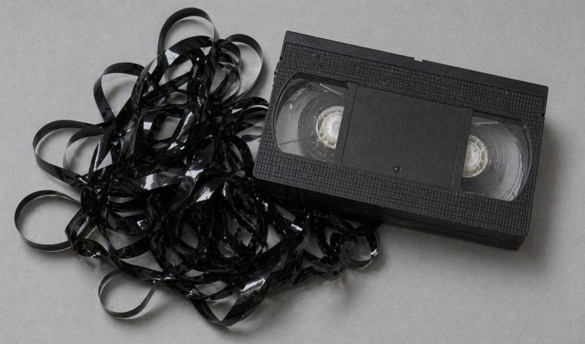 A VHS cassette beside a pile of tangled magnetic tape ribbon that has come loose from the internal spools, the kind of damage a stuck VCR can cause in seconds