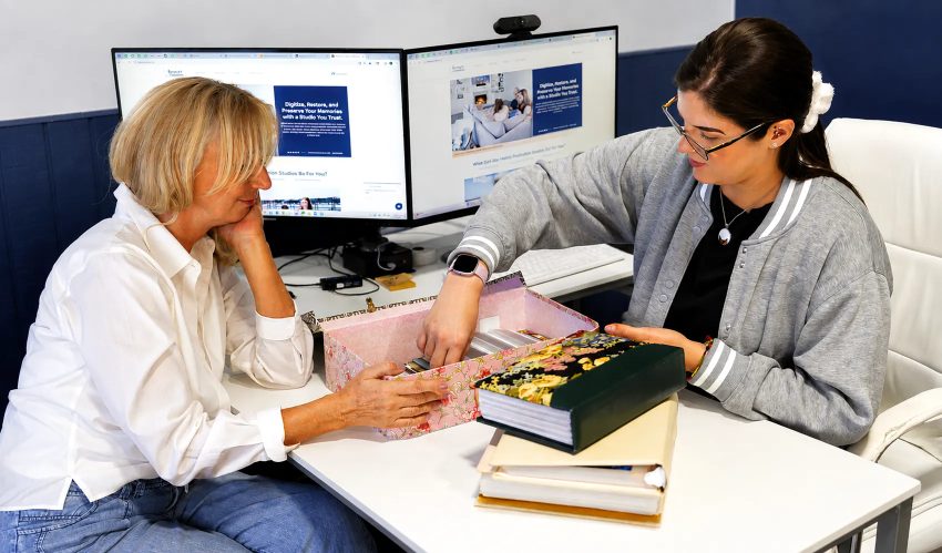 Two women reviewing printed photo albums at desk with dual monitors