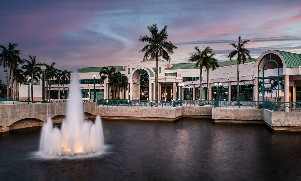 The Sunrise Civic Center behind a lake and fountain at dusk, with palm trees along the waterfront